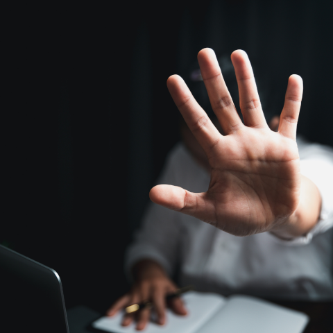 Person sitting at a desk in a dark room holding up their hand in a 'stop' gesture