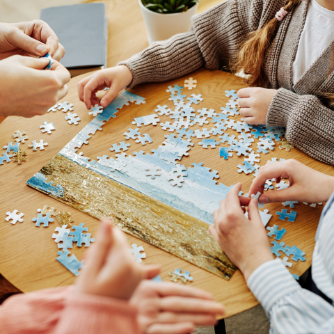 Photo of a group of people (only hands, partial arms inside the frame) putting together a jigsaw puzzle on a round table