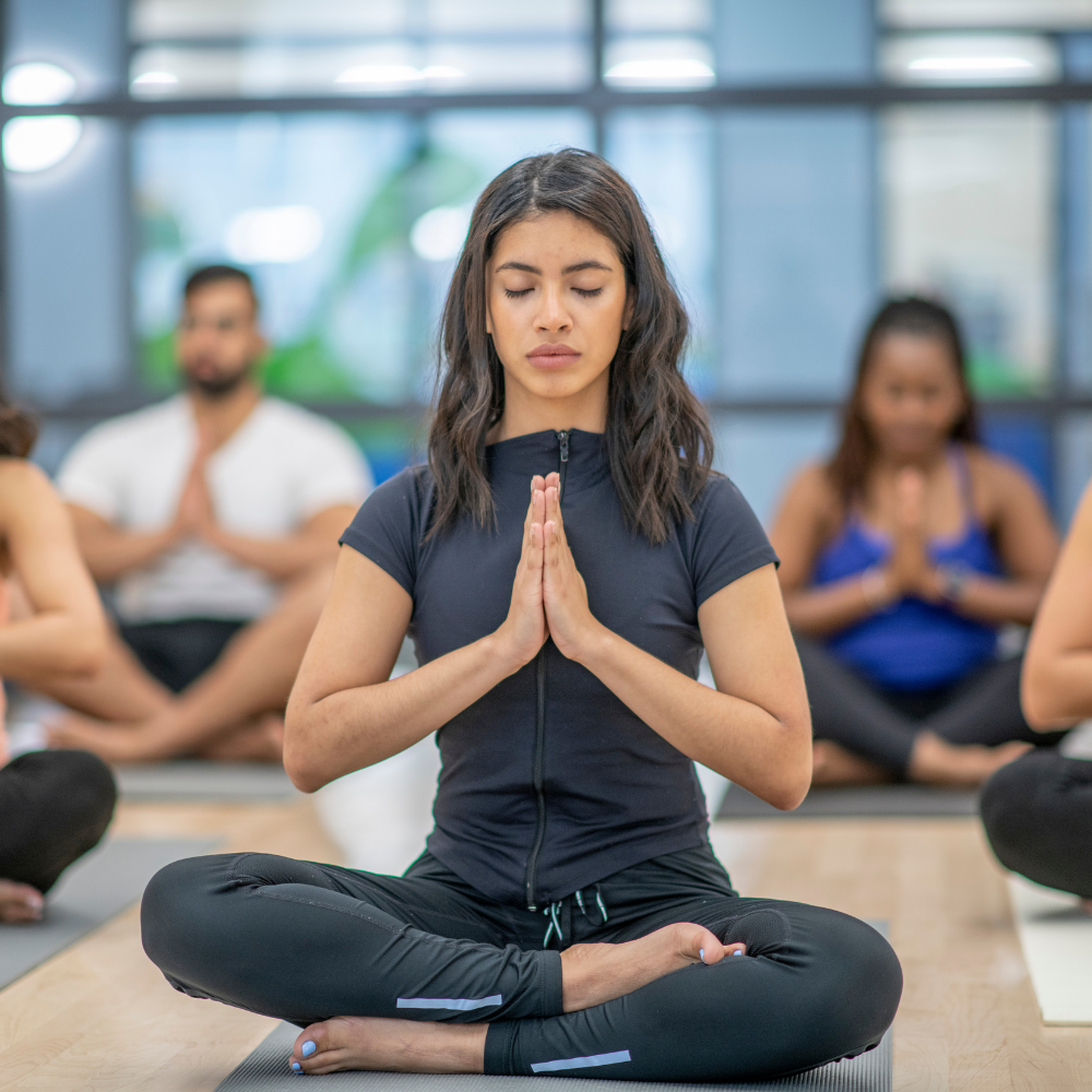 Photograph of individuals sitting crosslegged on the floor with their eyes closed and hands together at heart's center